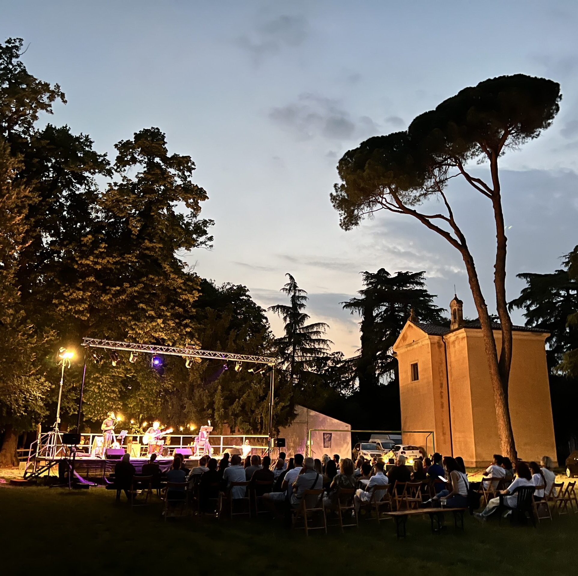 l'immagine ritrae un gruppo di persone sedute di fronte ad un palco all'aperto sul quale si sta svolgendo uno spettacolo con musica dal vivo, la foto è stata scattata al crepuscolo, con un grande albero che sovrasta il pubblico